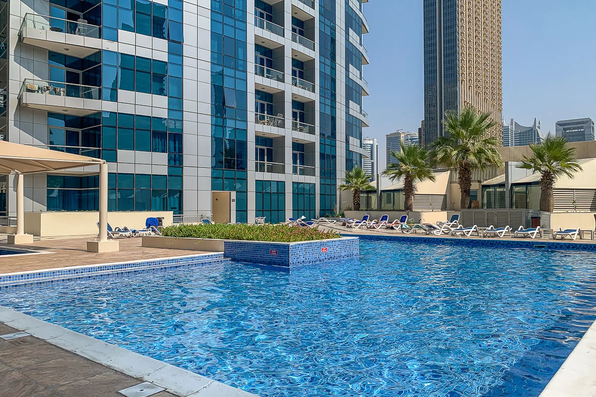Una piscina junto a edificios modernos, con palmeras y tumbonas alrededor.