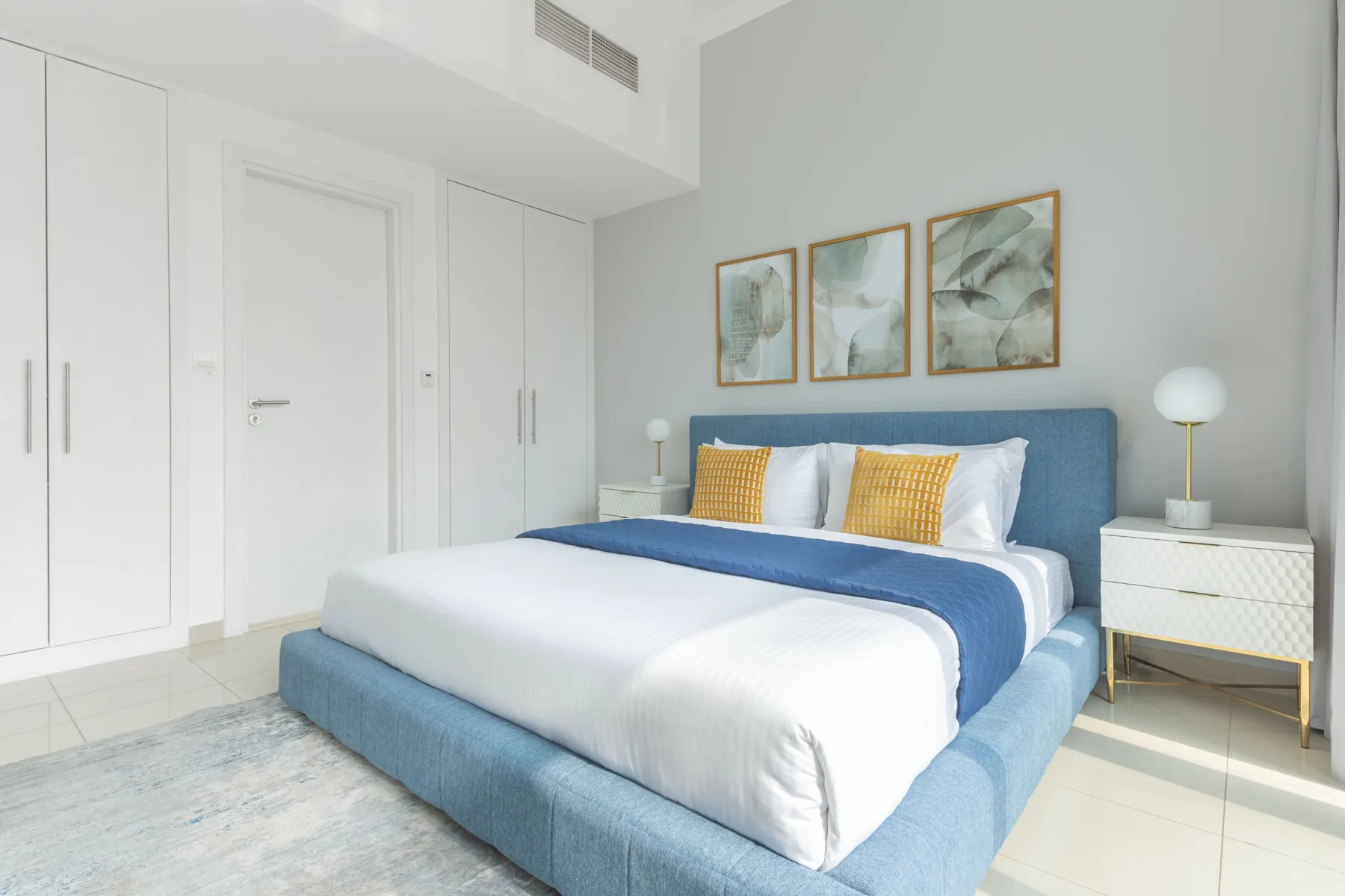 Un dormitorio luminoso con una cama azul, ropa de cama blanca y cuadros sobre el cabecero.
