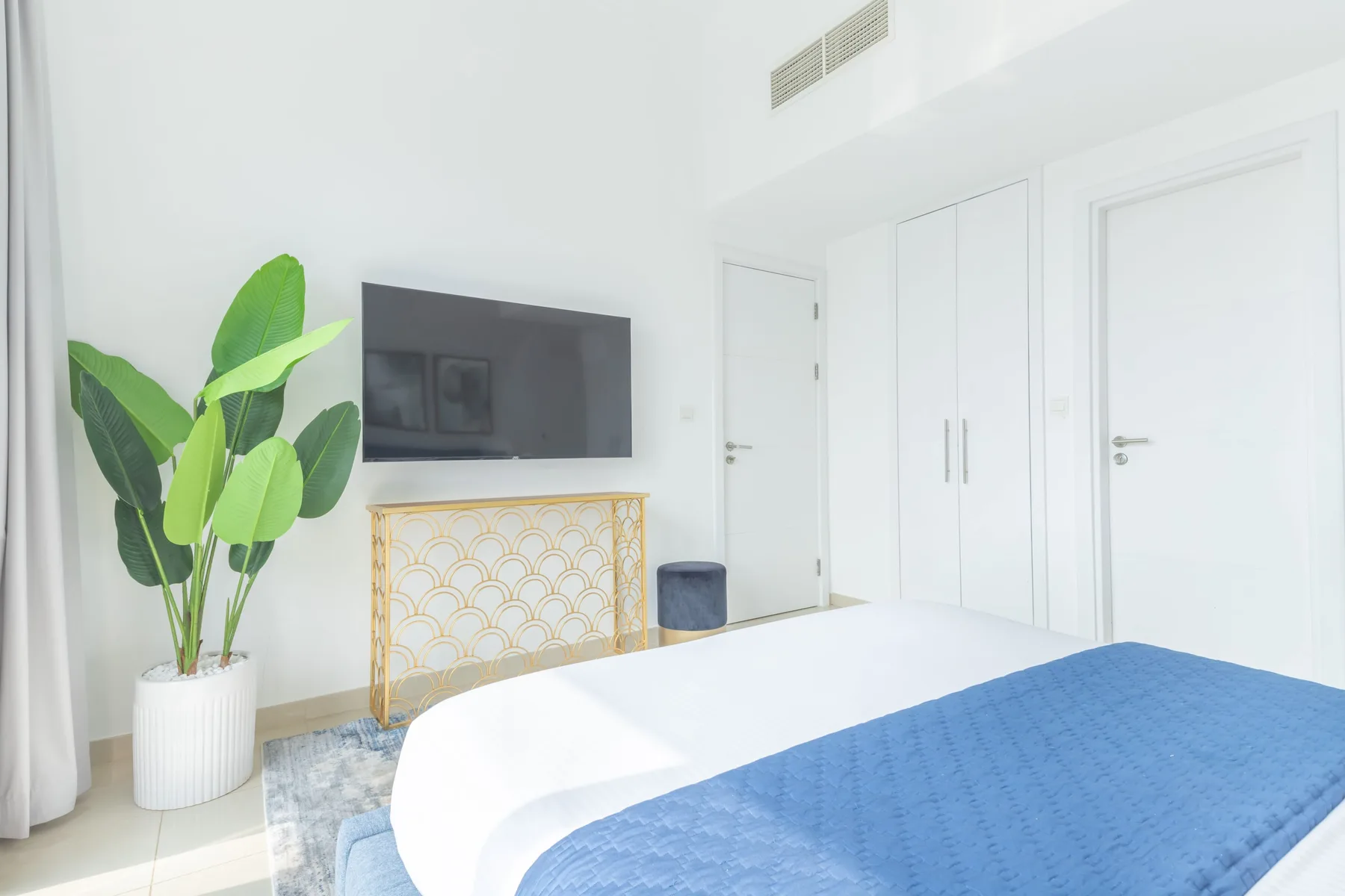 Un dormitorio luminoso con una cama, un televisor en la pared y una planta de hojas verdes.