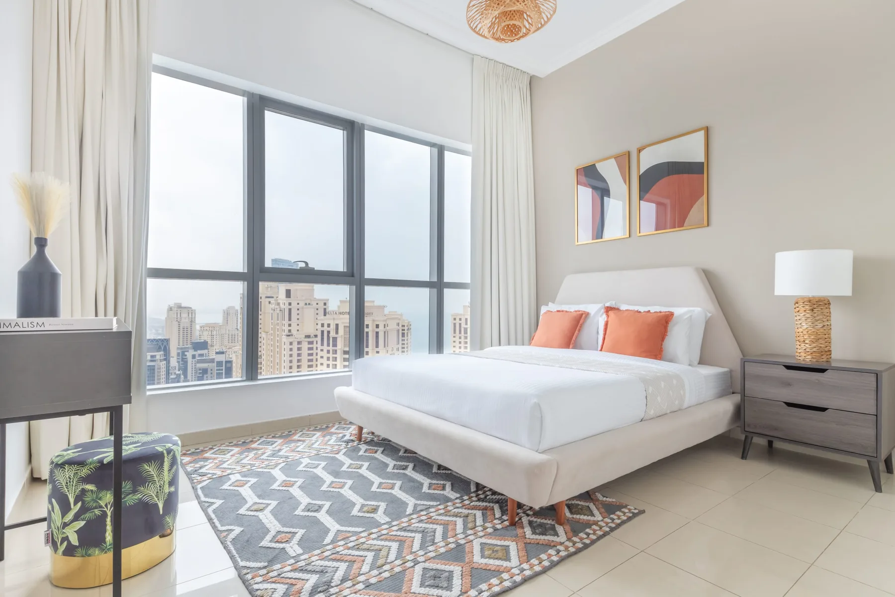 Un dormitorio luminoso con una cama blanca junto a grandes ventanas con vistas a edificios de la ciudad.
