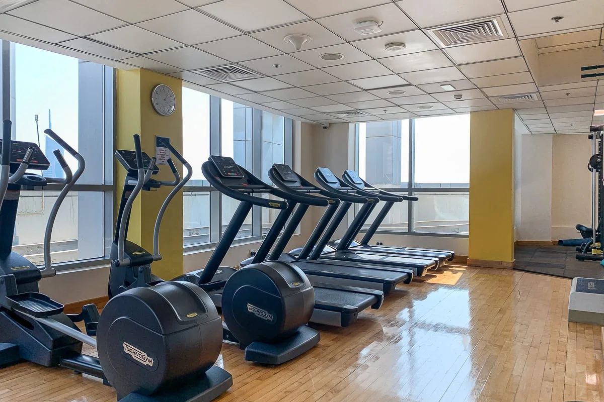 Un gimnasio con cintas de correr y máquinas de ejercicio junto a grandes ventanas.
