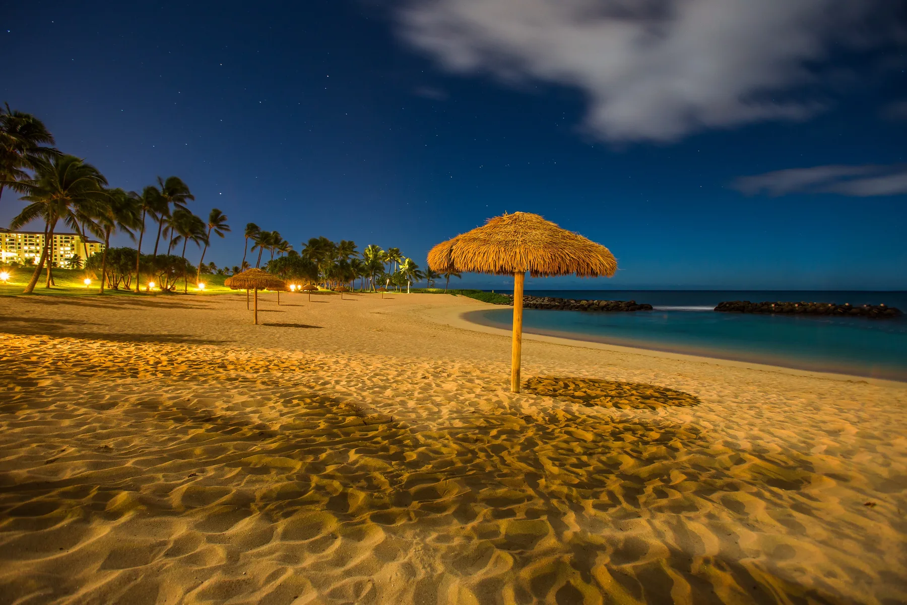 A sandy beach at night, with a thatched umbrella beside the water.