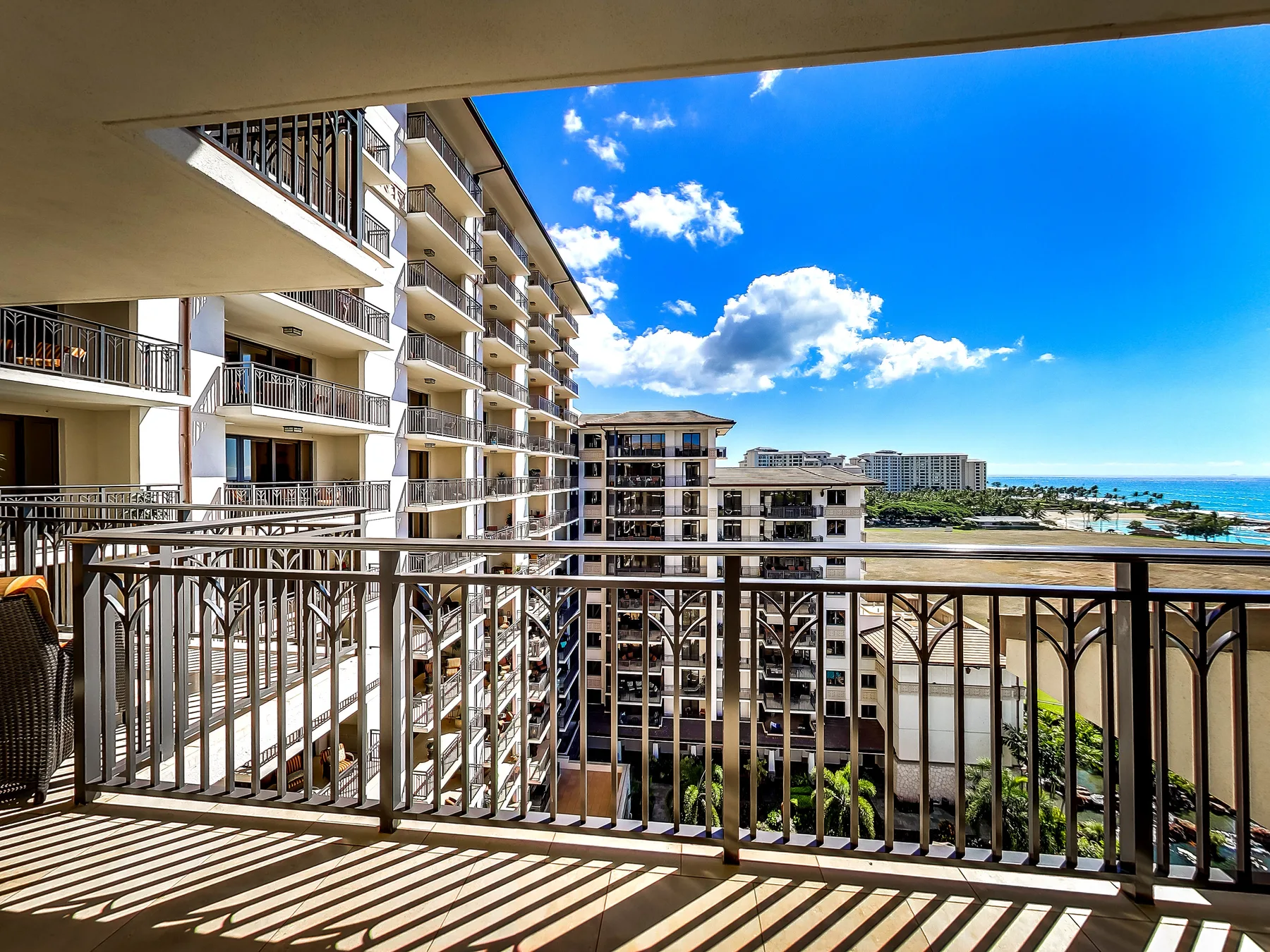 A balcony overlooks neighboring buildings and a coastal view beneath a bright blue sky.