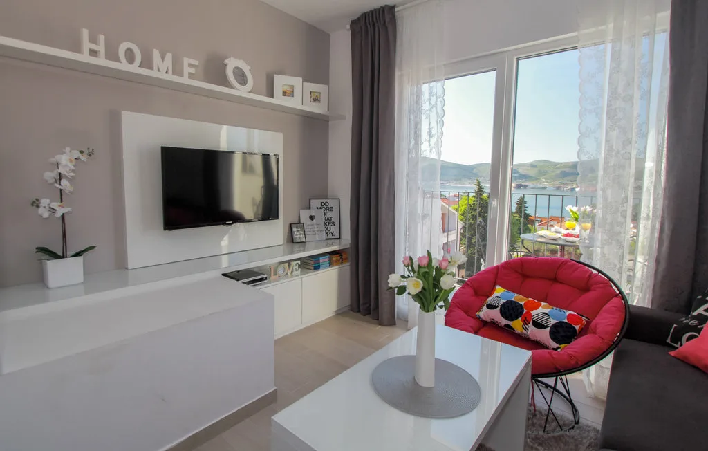 Una sala luminosa con un televisor montado en la pared y un sillón rojo redondo junto a las ventanas.