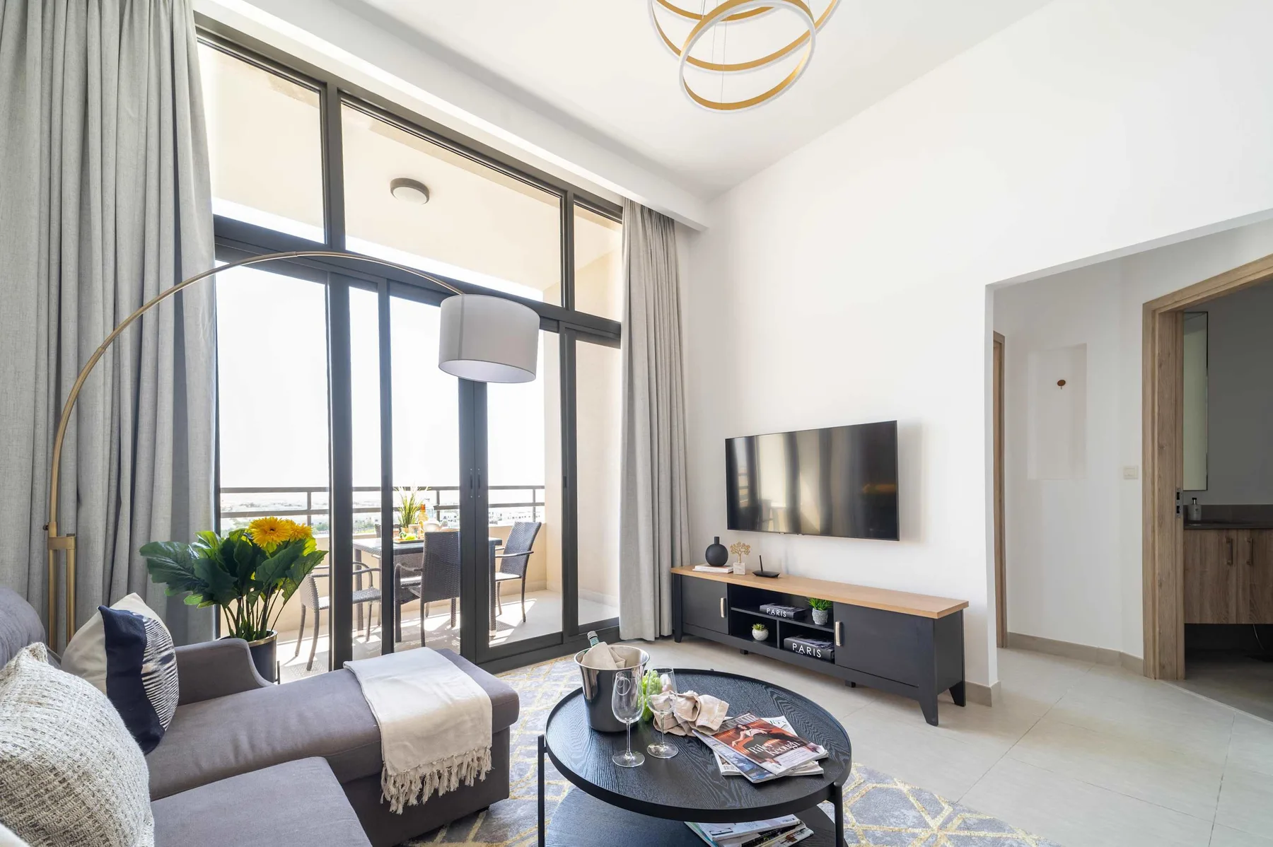Una sala luminosa con zona de estar, mesa de centro y televisor junto a ventanas altas.