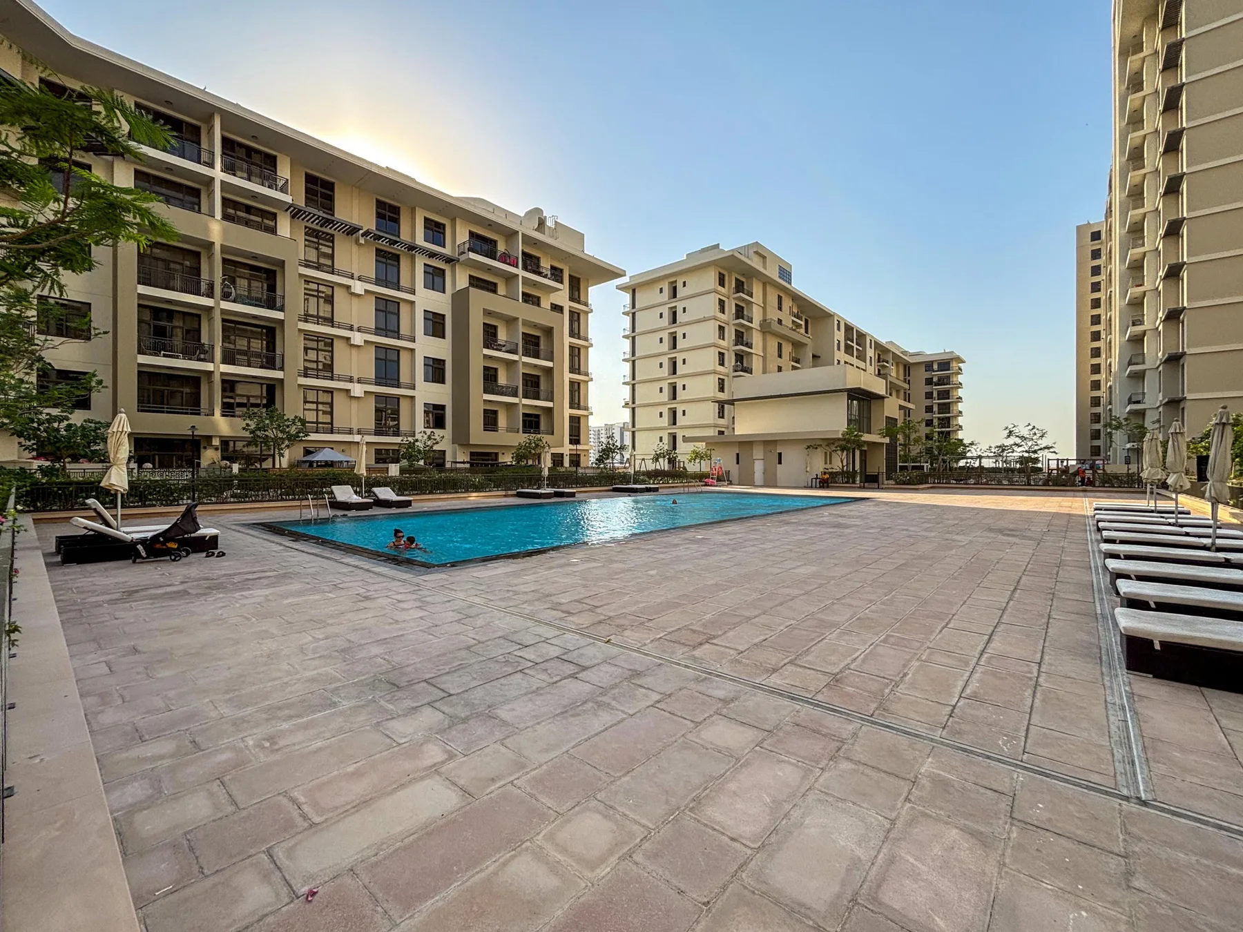 Una piscina al aire libre junto a una amplia terraza pavimentada y edificios de apartamentos.