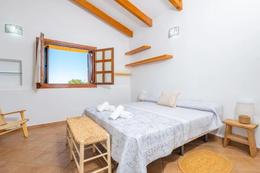 A bright bedroom with a bed, woven bench, and wooden ceiling beams.