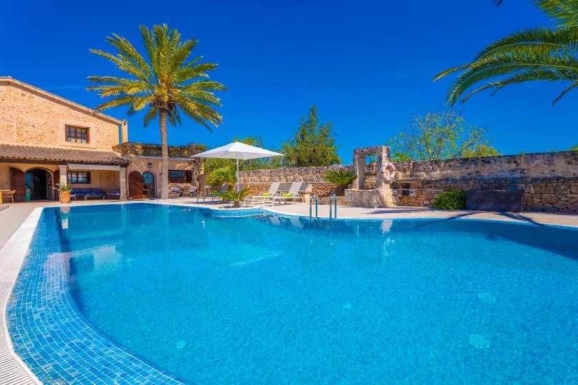 A swimming pool beside a stone building, with palm trees and an umbrella.