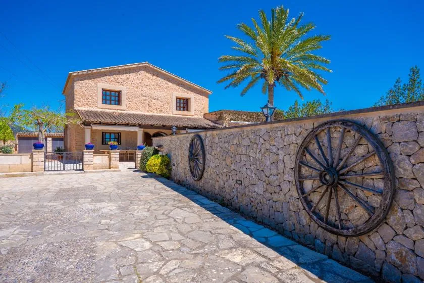 A stone-paved entrance leads to a pale stone building beside a wall with wooden wagon wheels.