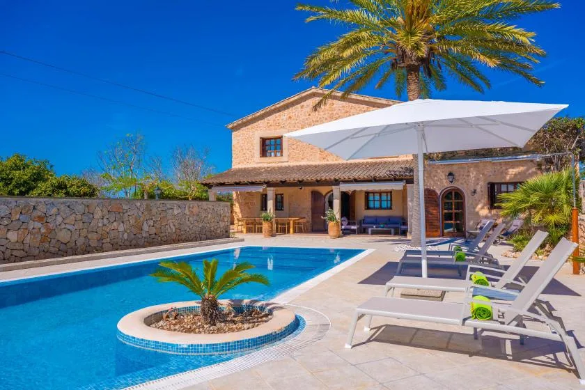 A sunny outdoor pool beside a stone house, with lounge seating and a large shade umbrella.