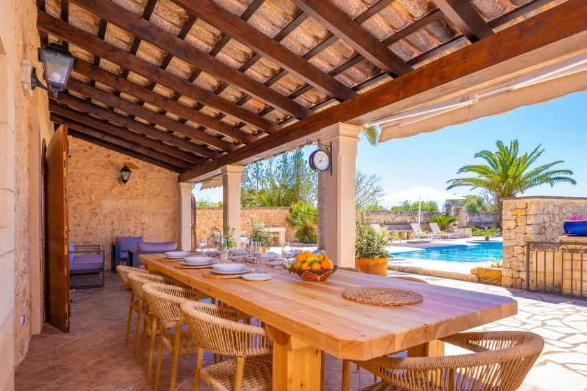 A covered outdoor dining area with a long wooden table beside a swimming pool.