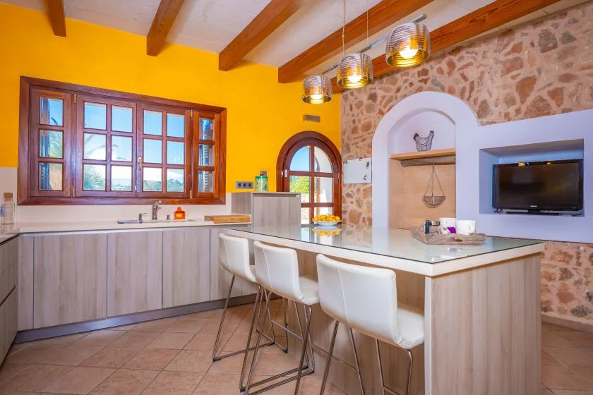A kitchen with a long counter, white seating, and a television set into the stone wall.