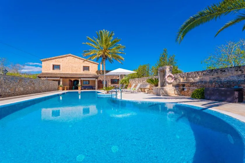 A swimming pool sits beside a stone house, with palm trees and lounge seating.