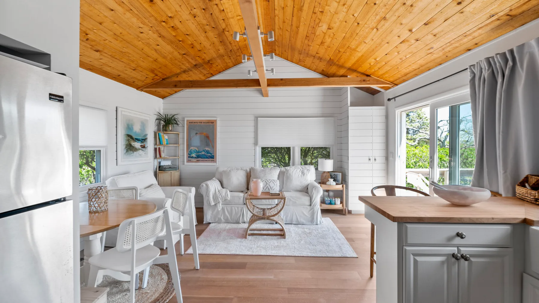 A bright living room opens into a kitchen, with seating beneath a wooden ceiling.