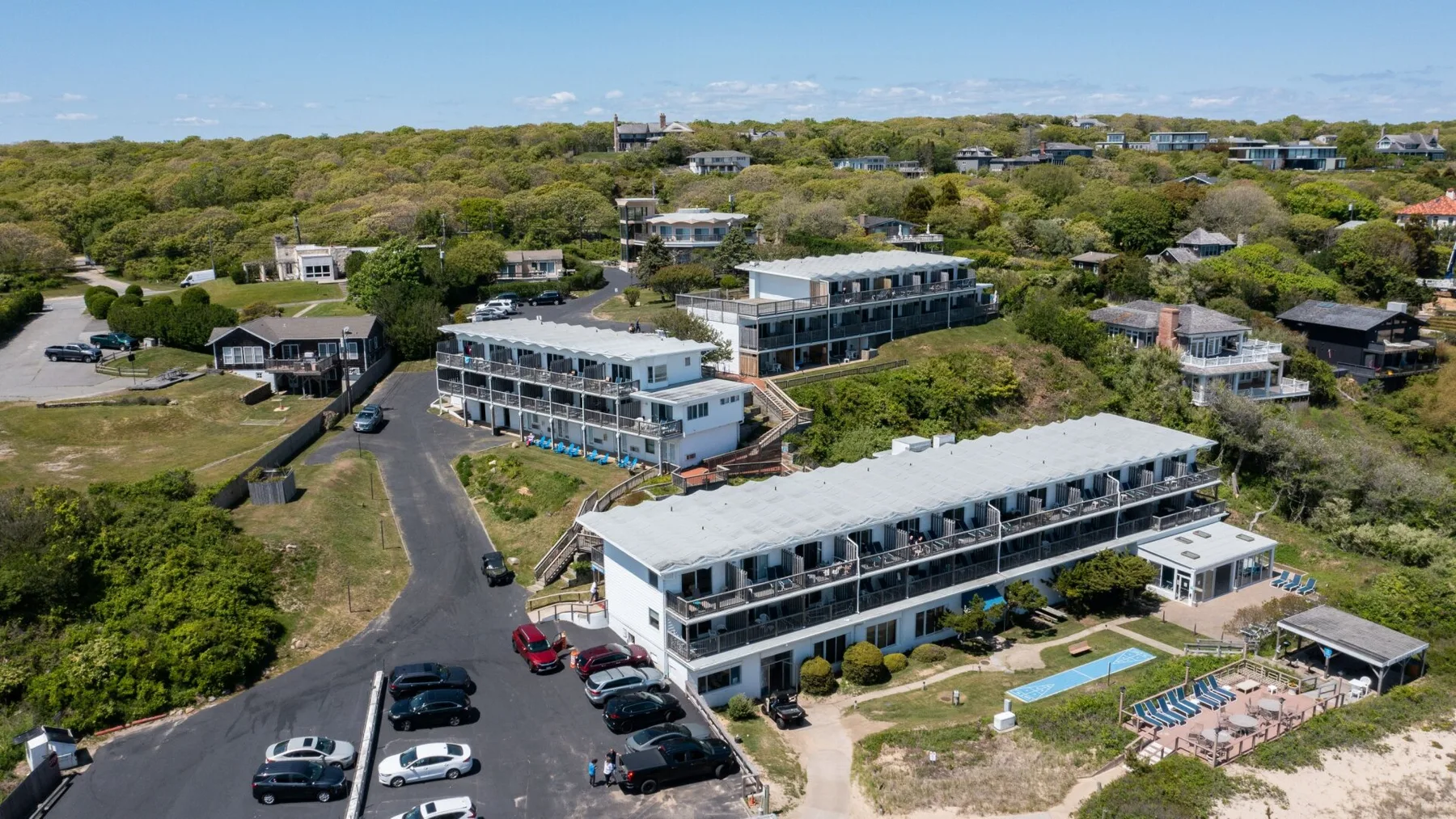 Aerial view of a coastal resort complex with long buildings, a pool, and surrounding greenery.