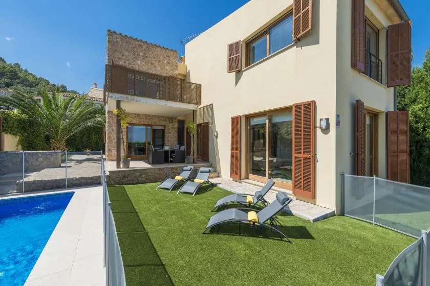 A modern house opens onto a grassy patio with lounge seating beside a swimming pool.