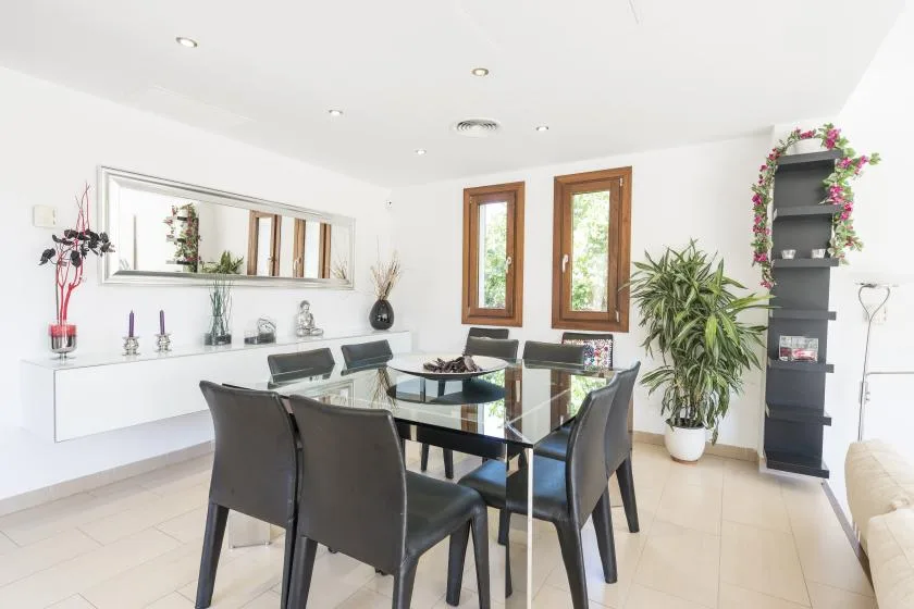 A bright dining area with a glass table, dark seating, and tall windows.