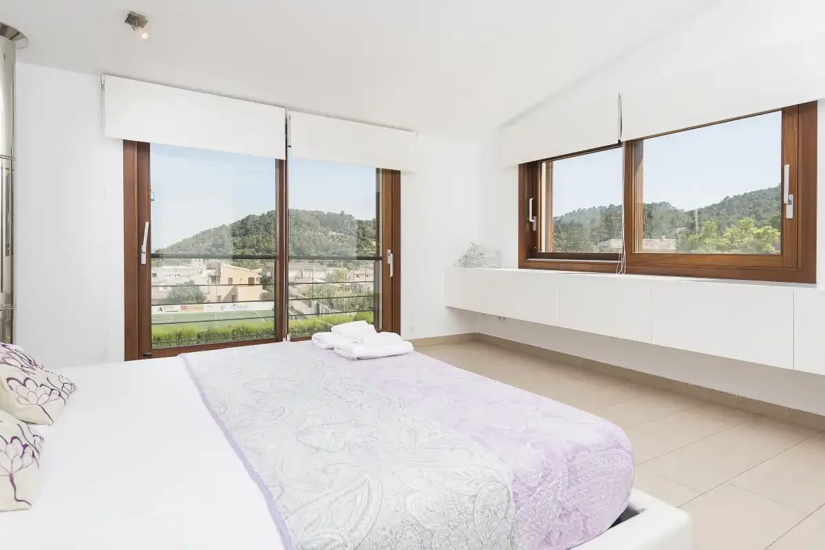 A bright bedroom with a bed beside large windows overlooking green hills.