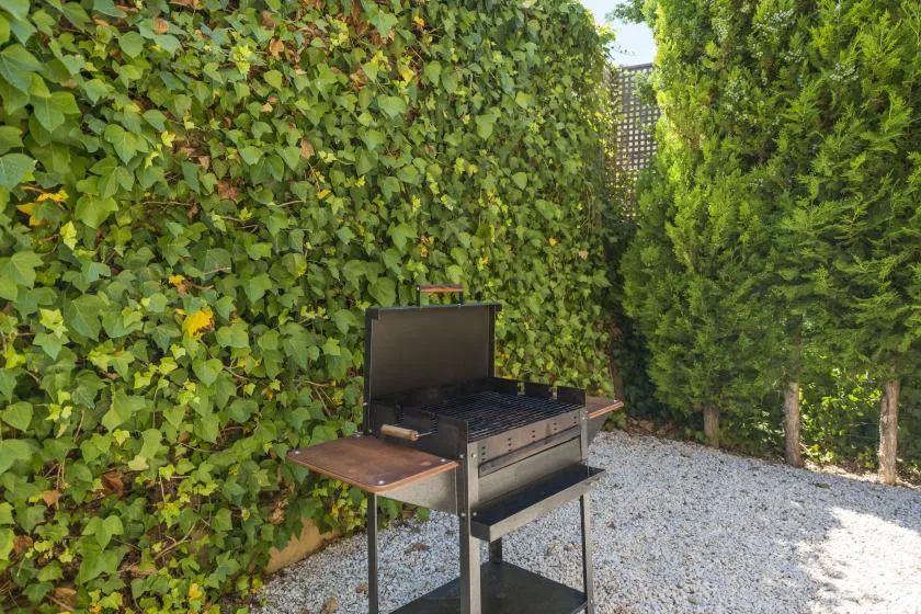 An outdoor grill stands beside leafy hedges on a gravel patio.