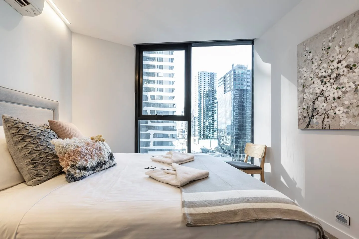 A bright bedroom features a bed beside a large window overlooking city buildings.