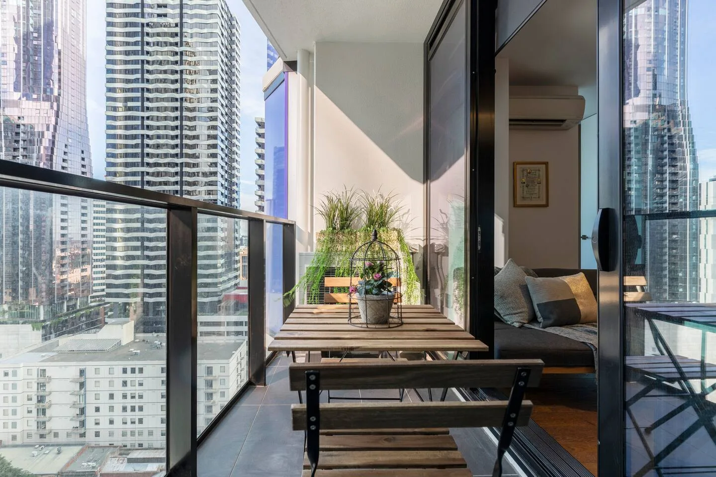 A furnished balcony with a narrow table and seating overlooks nearby high-rise buildings.