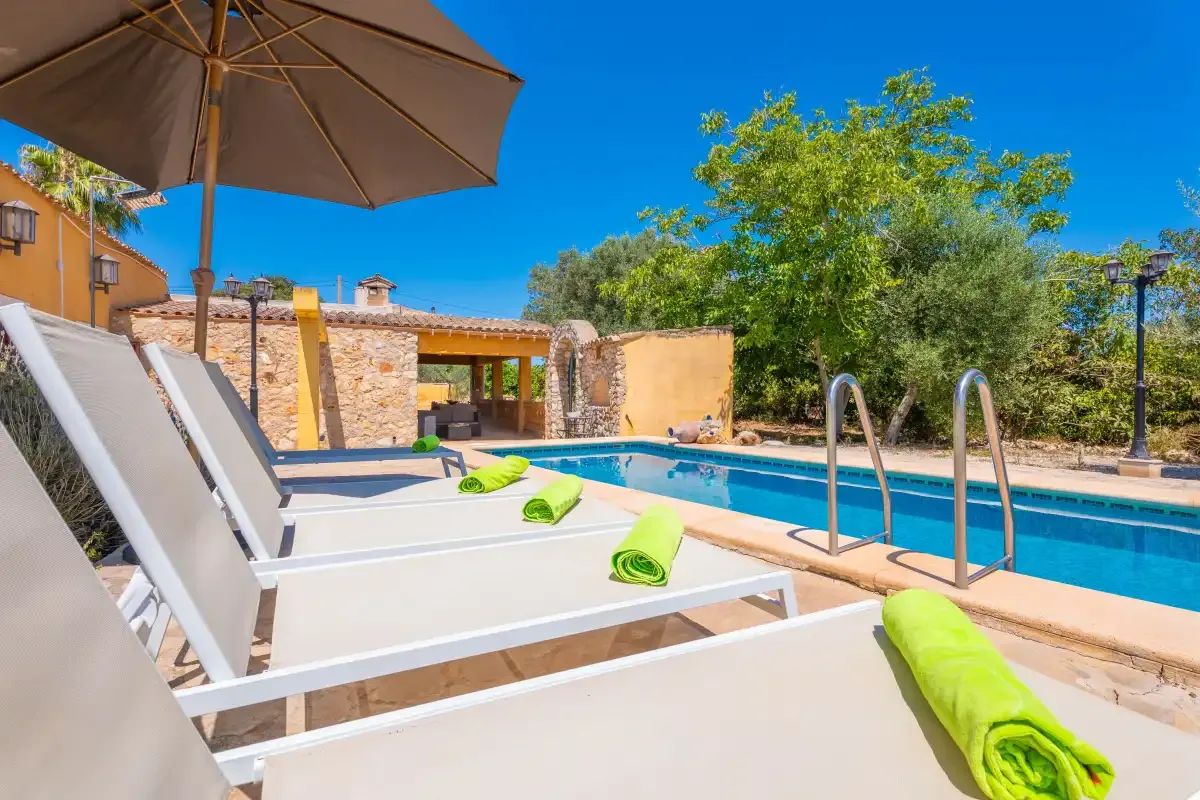 Una piscina al aire libre junto a un patio soleado con tumbonas y una sombrilla grande.