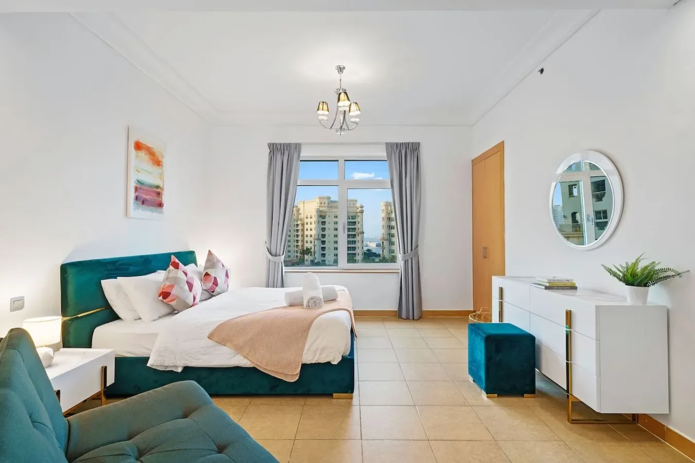 A bright bedroom with a bed, bedside seating, and a window overlooking nearby buildings.