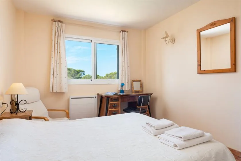 A bedroom with a desk beneath a window and folded towels on the bed.