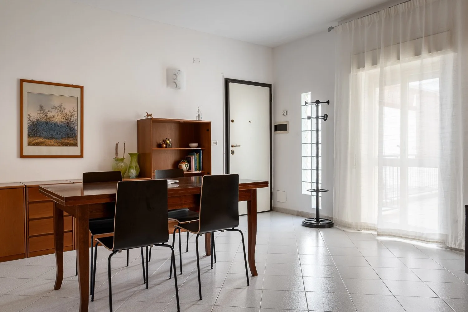 Un comedor luminoso con una mesa de madera, sillas oscuras y cortinas translúcidas.