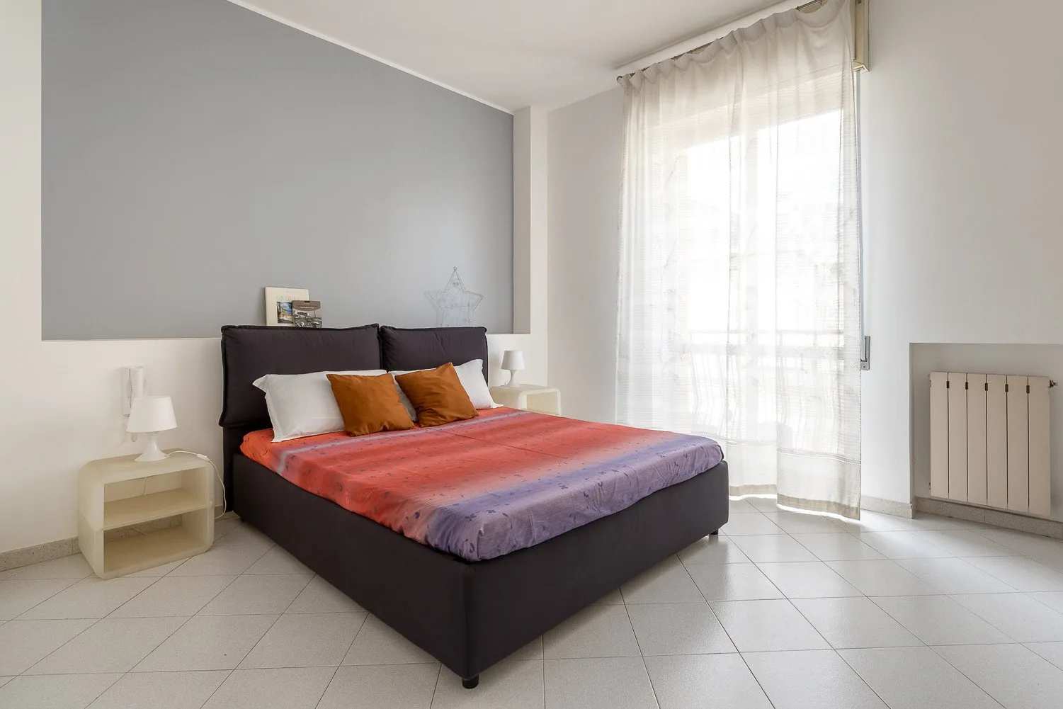 Un dormitorio con una cama vestida con ropa de cama coral y morada junto a una ventana luminosa.