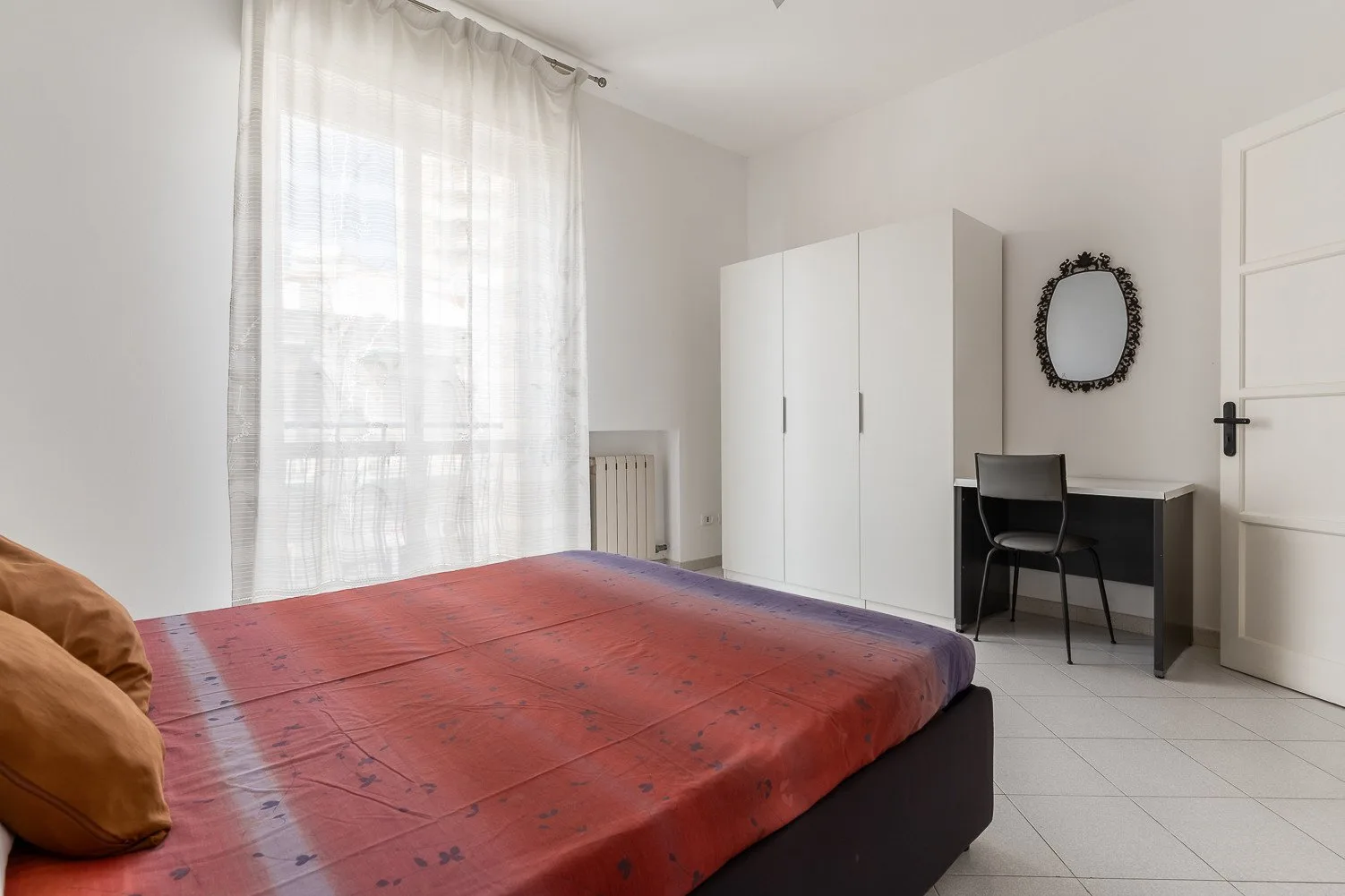Un dormitorio con una cama cubierta de rojo, armario y escritorio junto a una ventana con cortinas.