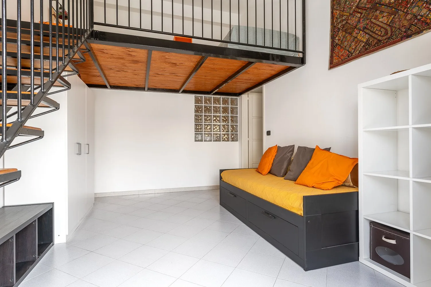 Una habitación luminosa con una plataforma elevada para dormir y un diván con cojines naranjas.