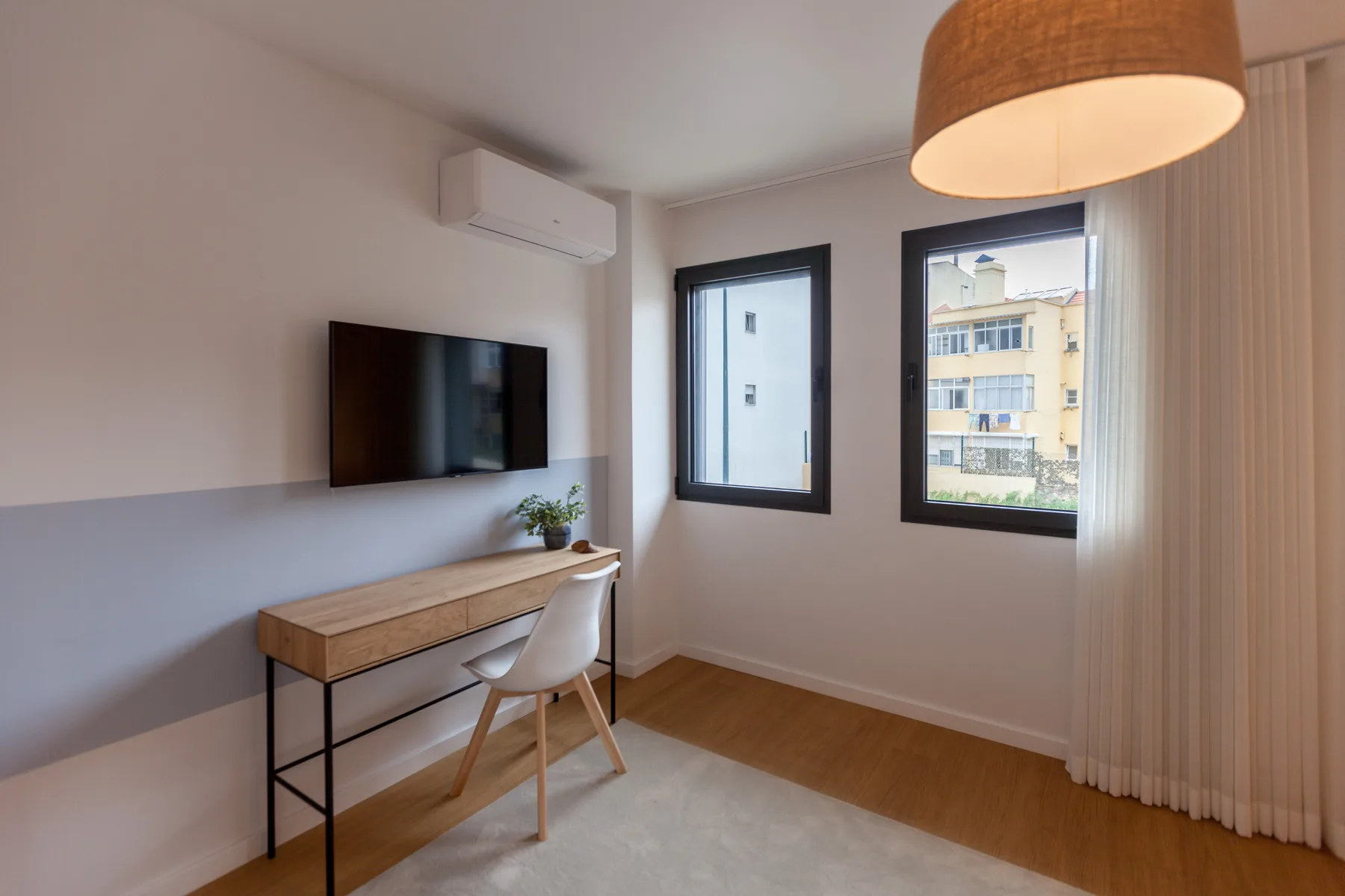 Una habitación luminosa con escritorio, asiento y ventanas con vistas a edificios cercanos.