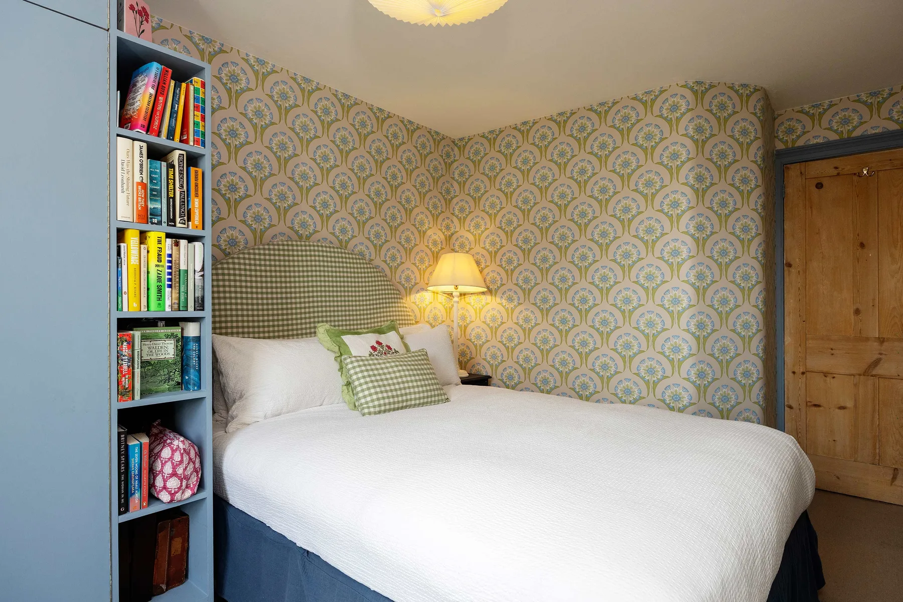 Un dormitorio con una cama bien tendida, papel pintado estampado y una estantería estrecha.