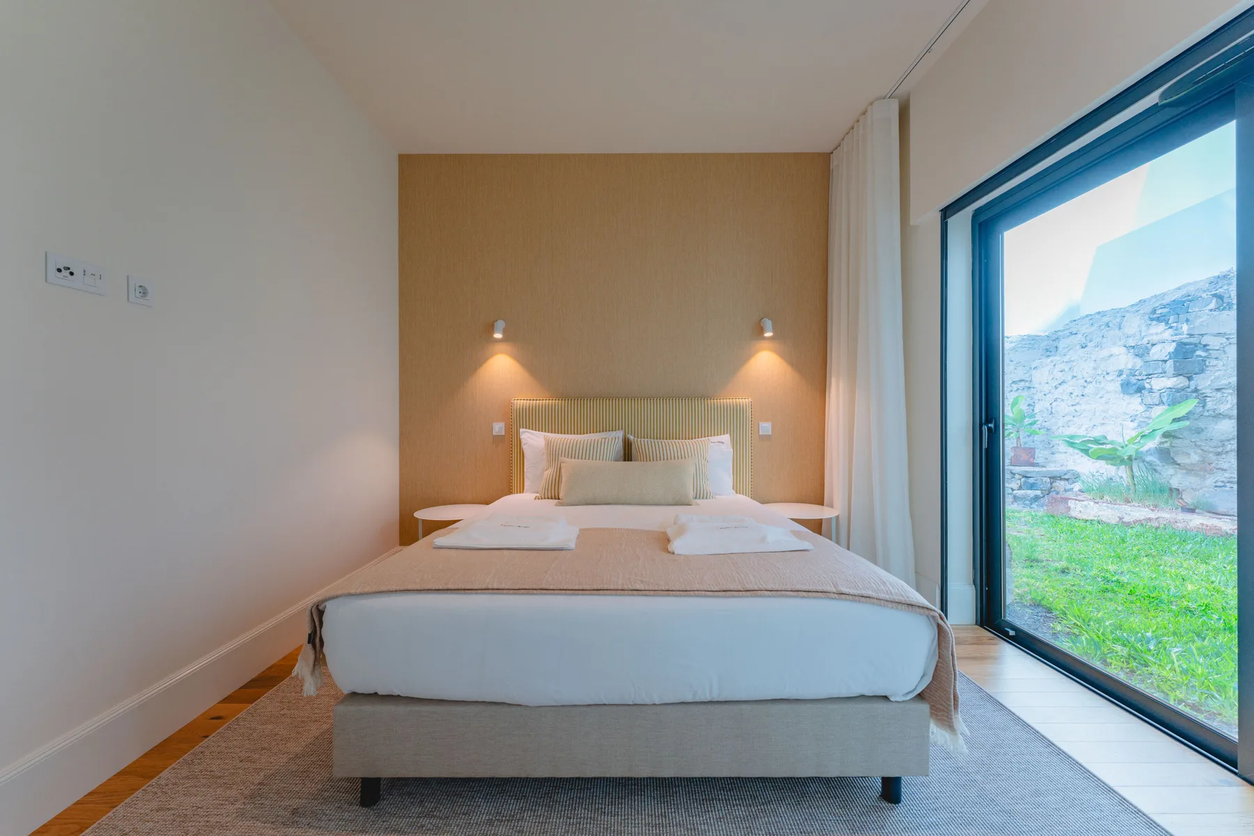 A bedroom with a neatly made bed and a large window overlooking greenery.