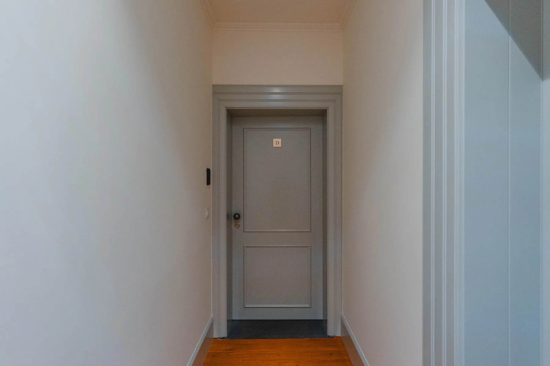 A narrow interior hallway leads to a closed door, with wood flooring.