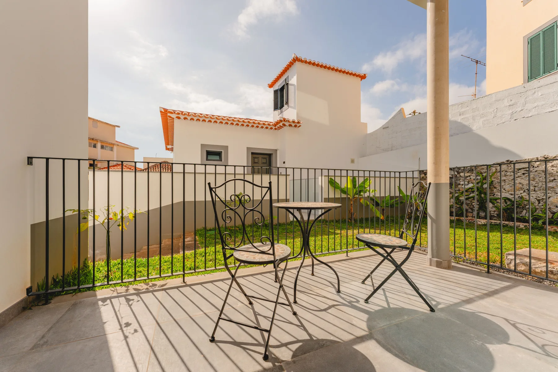 A sunny balcony with metal seating overlooks a small garden and nearby buildings.