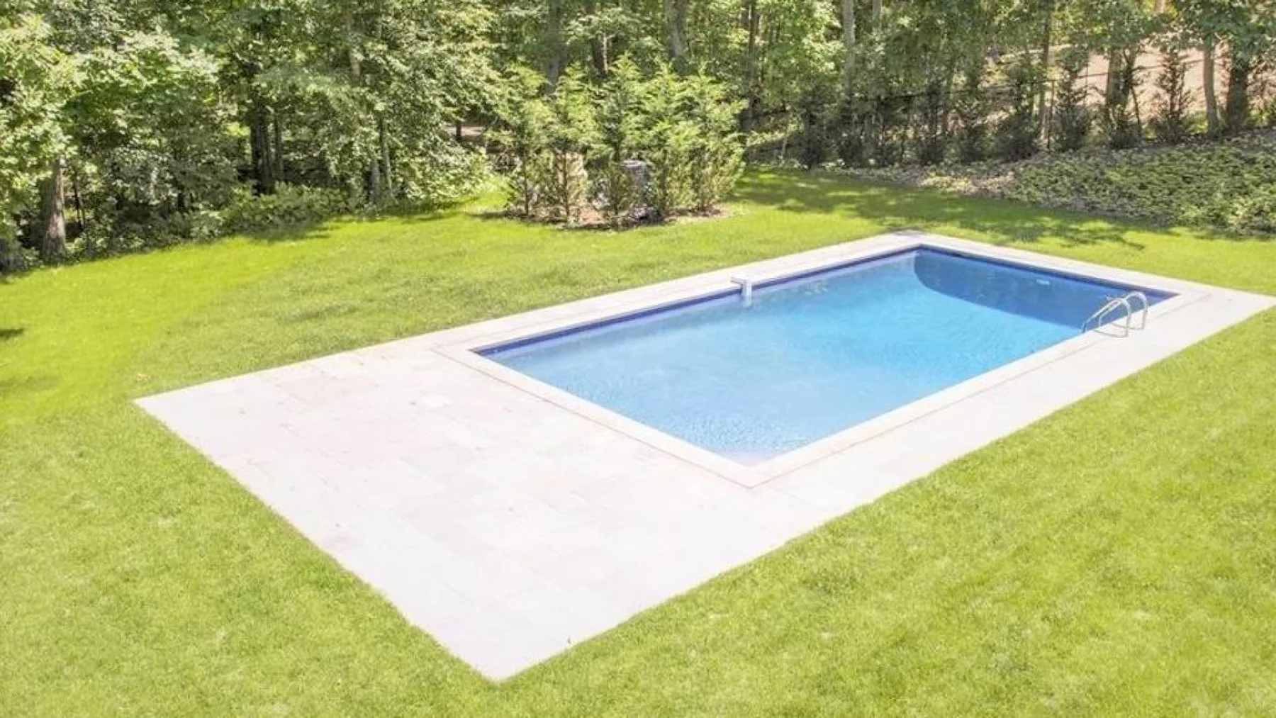 An outdoor swimming pool sits in a grassy yard bordered by trees.