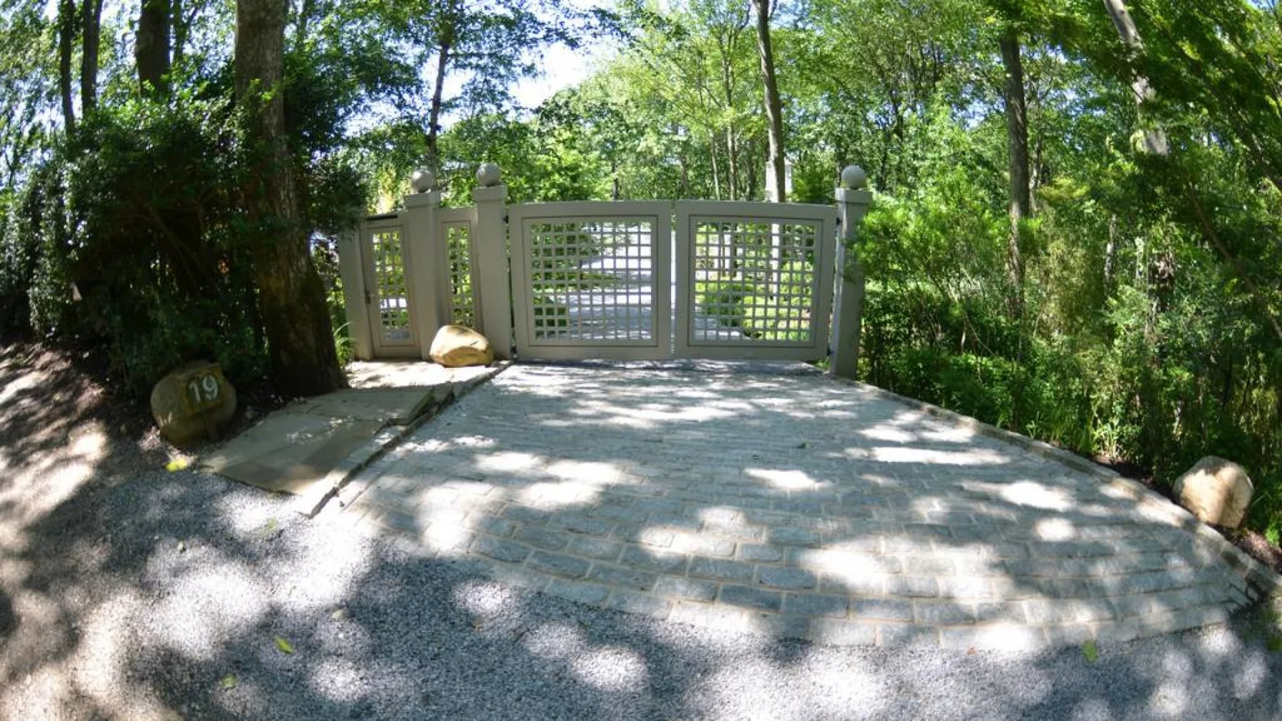 A shaded outdoor driveway leads to a gated entrance amid trees and greenery.