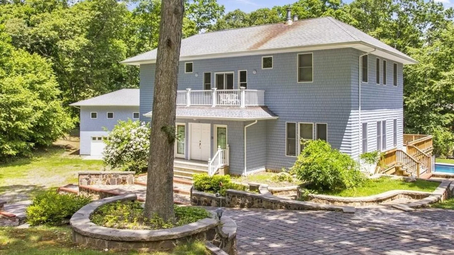 A gray two-story house with a balcony stands among trees beside a paved driveway.