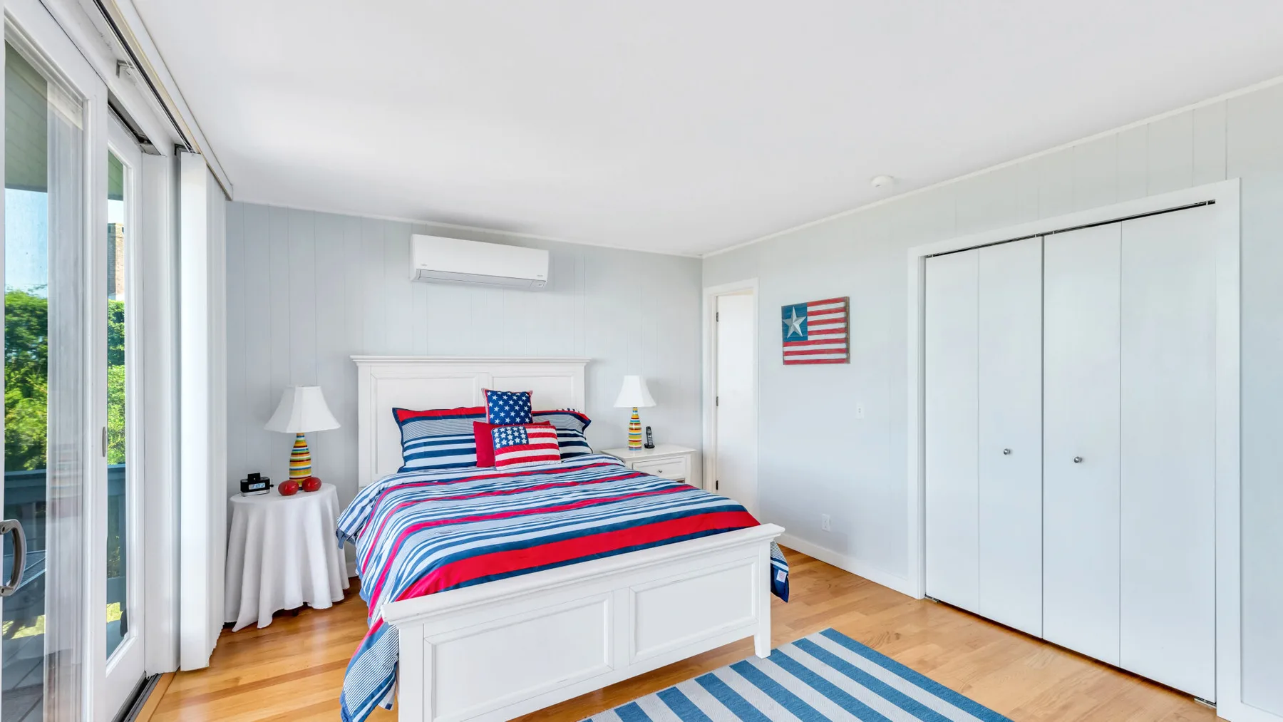 Un dormitorio luminoso con una cama vestida con ropa de cama roja, blanca y azul.