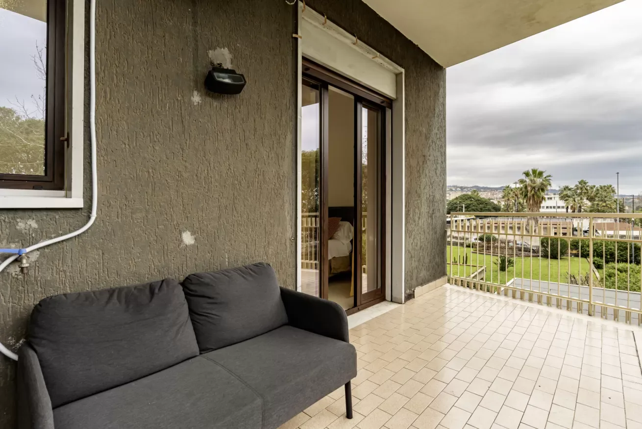 Un balcón cubierto con asientos oscuros y puertas corredizas de vidrio da a una zona ajardinada.