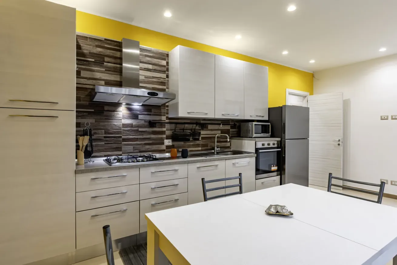 Una cocina luminosa con armarios blancos, zona de cocción y una mesa grande.
