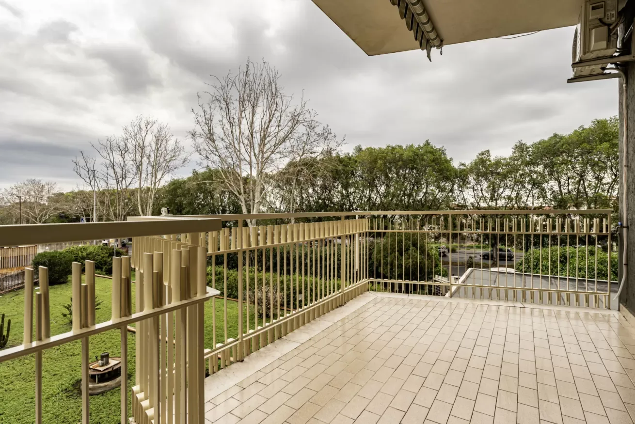 Una terraza exterior embaldosada da a un jardín frondoso detrás de una barandilla metálica.