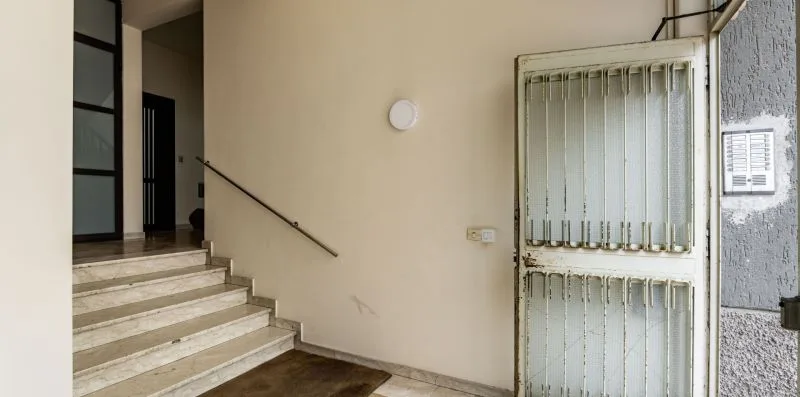 Una escalera interior con peldaños claros, un pasamanos en la pared y una reja metálica.