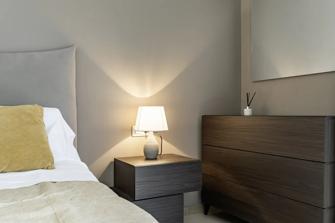 Un dormitorio con una cama arreglada, una mesita de noche y una lámpara encendida.