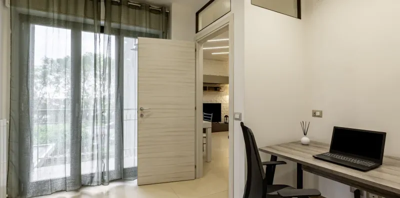Un escritorio con una computadora portátil abierta está junto a una puerta y puertas de vidrio con cortinas.