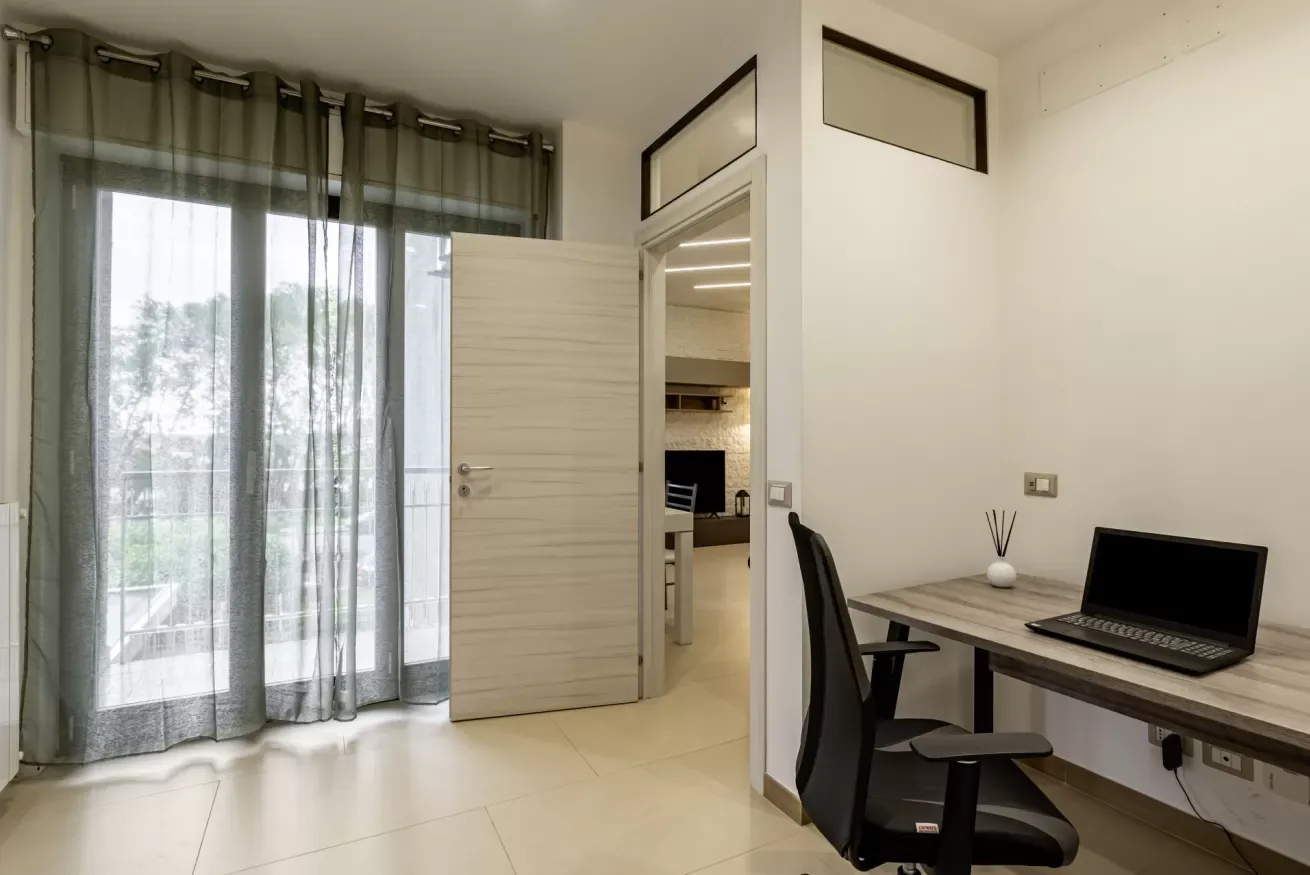 Un pequeño espacio de trabajo con escritorio y silla junto a una puerta de vidrio con cortinas.