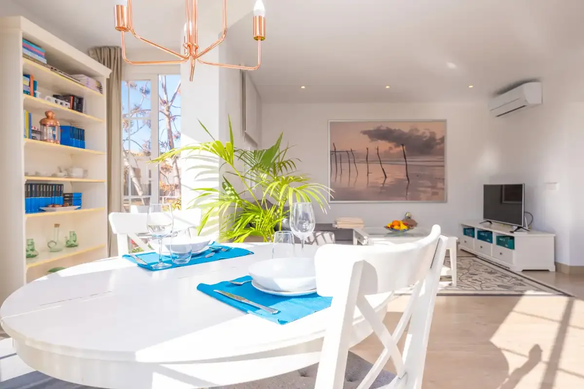 A bright dining area with a set table, shelving, and a living space beyond.