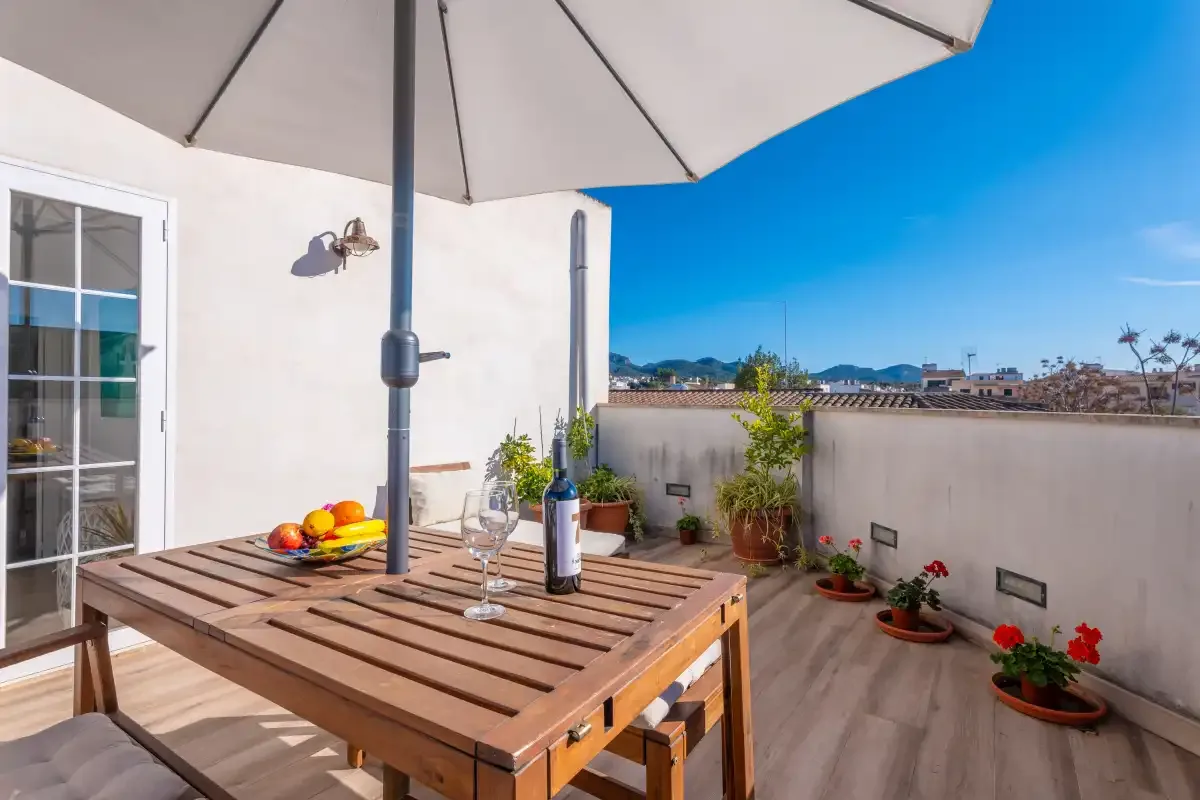 A sunny rooftop terrace with a wooden dining table, umbrella, and potted plants.